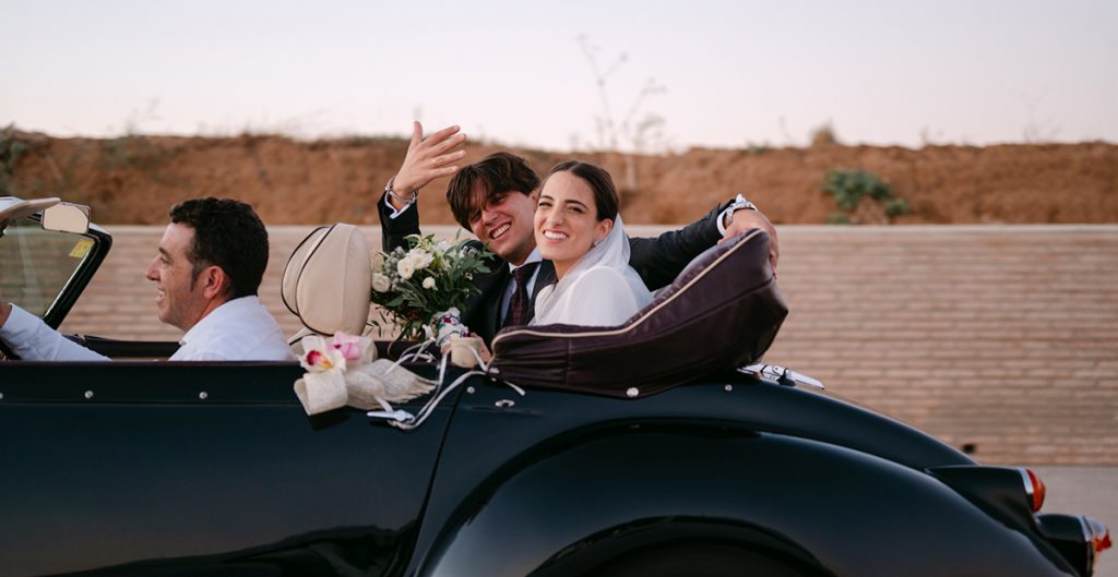 Fotografía divertida de boda novios saludando desde el coche al fotografo de boda